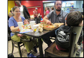 Picture shows a family sitting at a table in a food court.  The mother is holding a baby, and there is a little boy with a T-shirt displaying robotics on his back.  His father is getting some of the pizza that's on the table, and reaching out to the little boy.